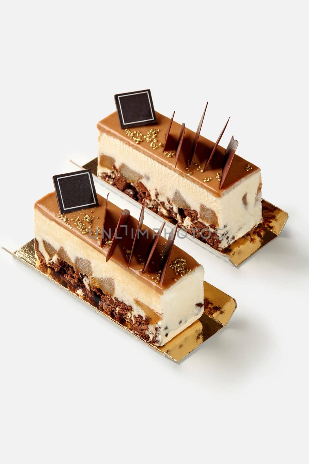 Two appetizing creamy mousse desserts with crunchy streusel, pieces of spiced poached pears and soft caramel topping decorated with dark chocolate flakes and golden sugar sprinkles, isolated on white