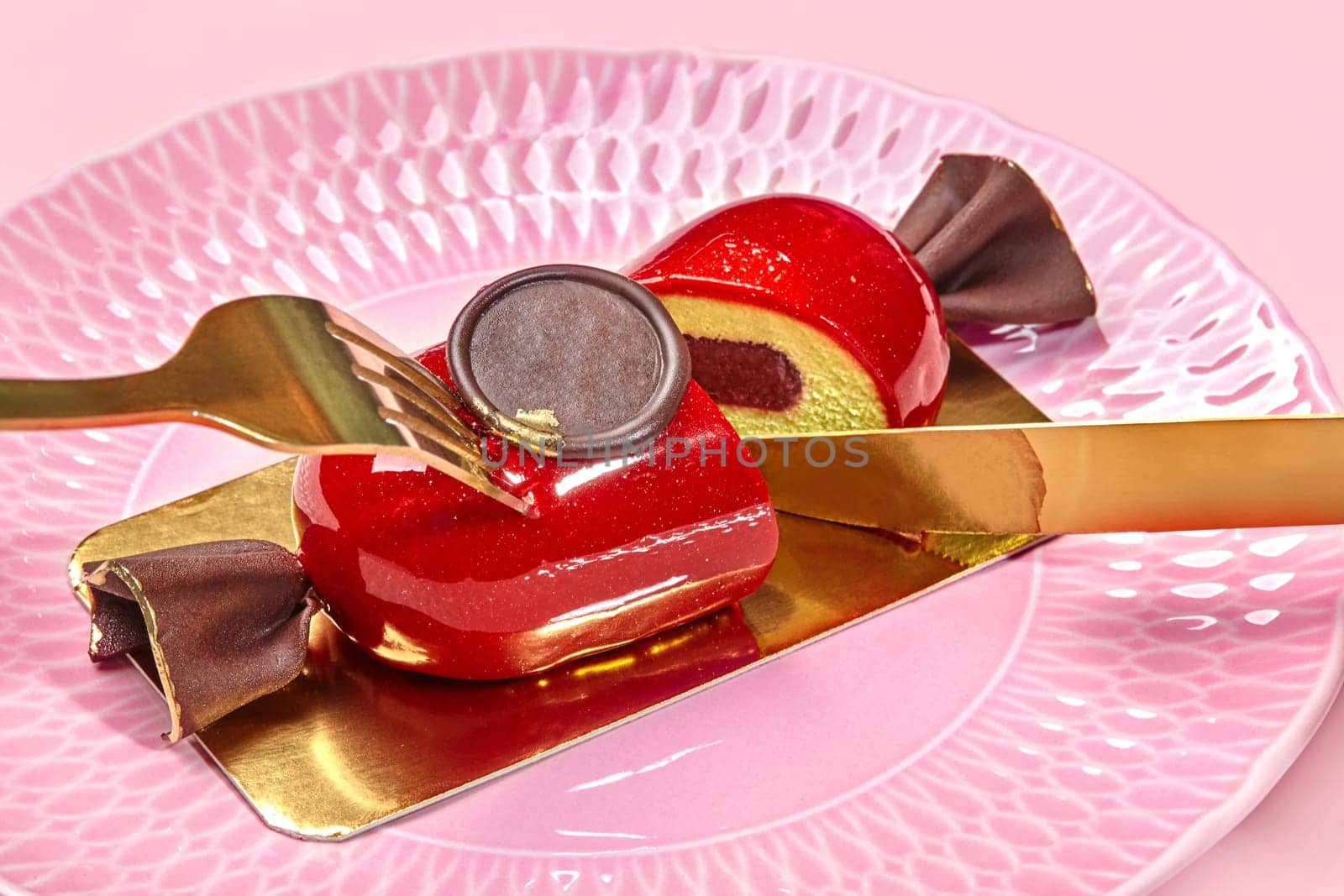 Close-up of elegant gold cutlery slicing candy-shaped mousse dessert with berry jelly and glossy red glaze served on textured pink plate