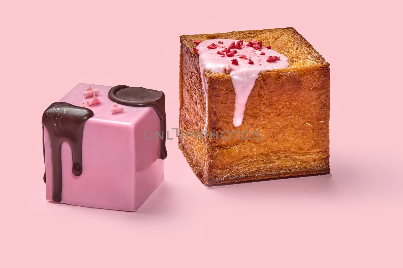 Two pastry cube croissants dripping with chocolate and creamy berry glaze and freeze-dried raspberry pieces, against vibrant pink background. Modern confectionery in classic French tradition