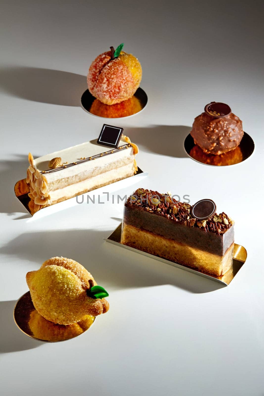 Variety of handcrafted desserts, velvety mousse and sponge layered cakes, crispy shortcrust pastries dusted with sugar in shape of fruits and chocolate praline truffle spotlighted on light surface