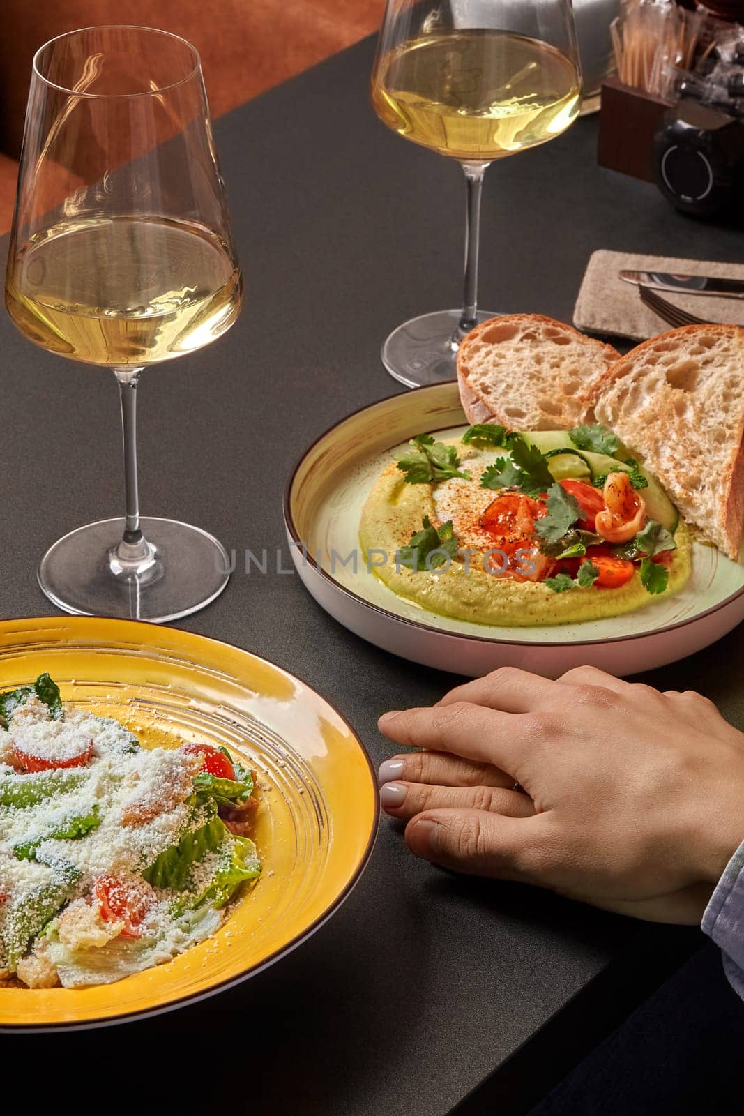 Tender moment of connection with hands intertwined over romantic dinner of fresh Caesar salad with grated Parmesan and creamy hummus topped with herbs and shrimps, accompanied by white wine glasses