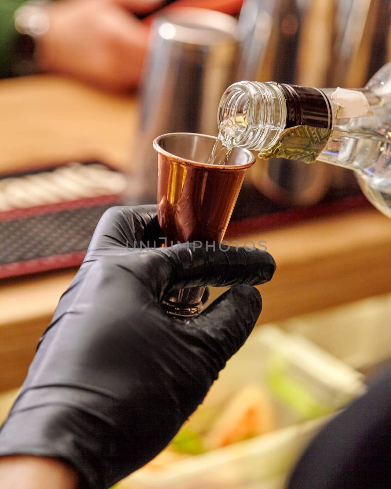 Bartender hand in black glove expertly pouring clear alcohol drink from bottle into gleaming copper jigger, measuring out spirits to ensure perfect balance for cocktail