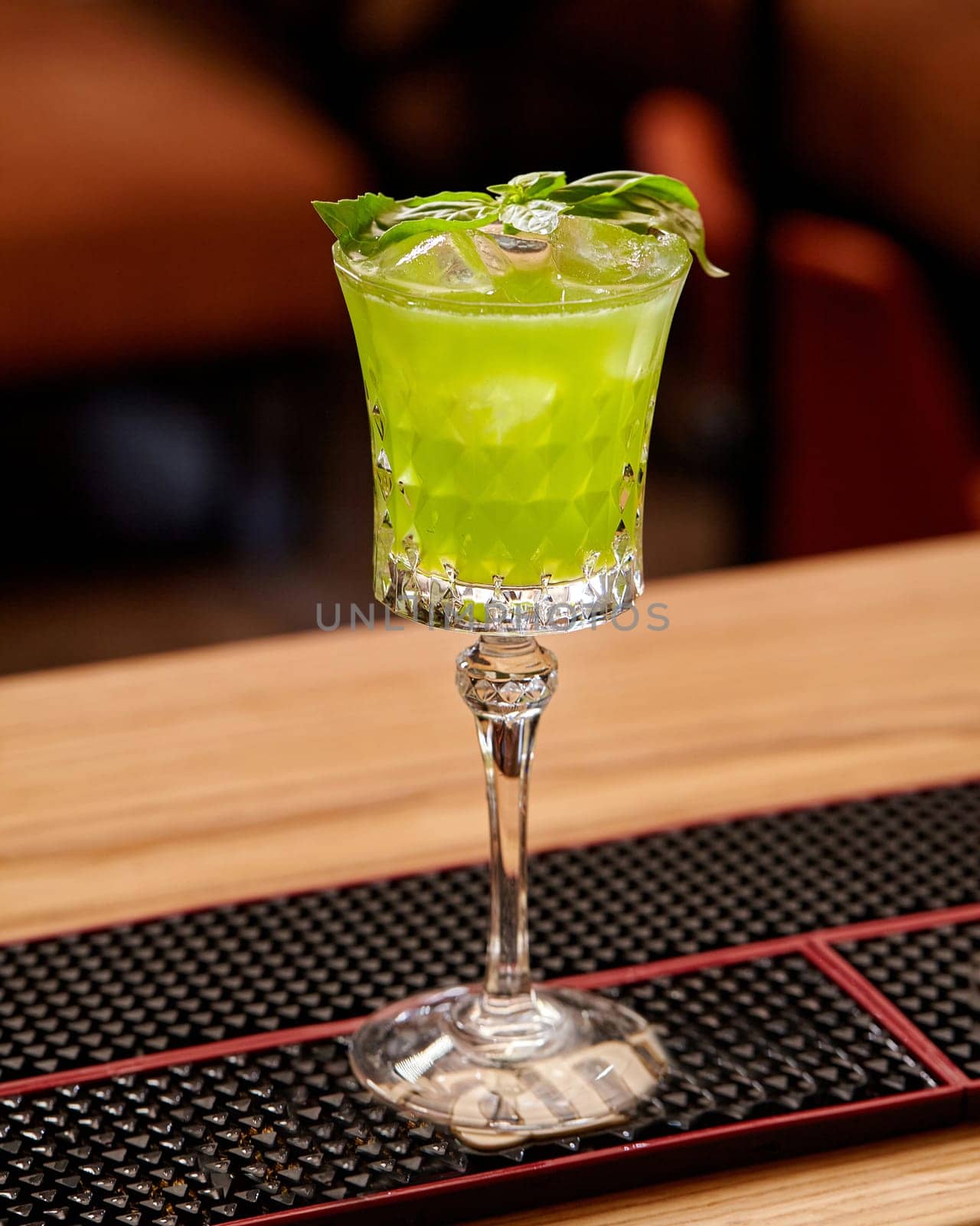 Glass of chilled gin based Basil Smash cocktail with syrup, lemon juice and ice cubes garnished with fresh green basil leaves, poised on bar mat