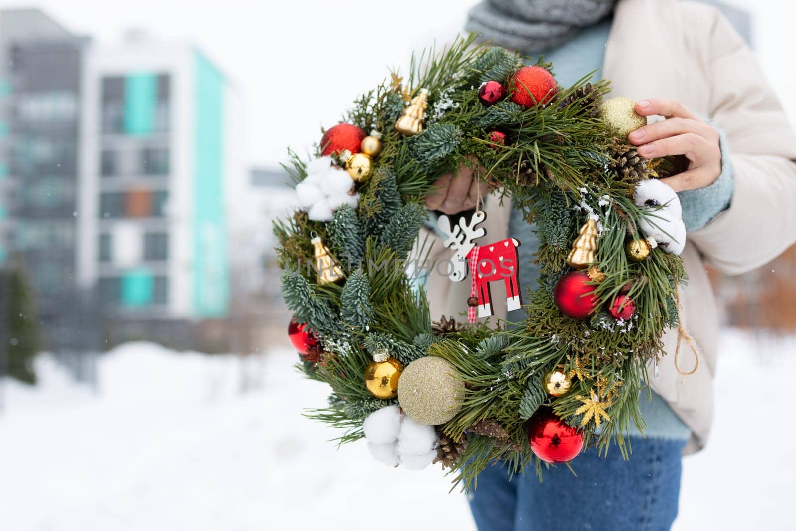 A woman is standing in the snow, holding a Christmas wreath. The snow-covered ground contrasts with the green of the wreath, creating a festive scene.