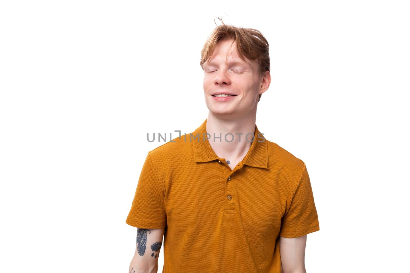 a young red-haired guy in an orange t-shirt shyly closed his eyes on a white background with copy space.