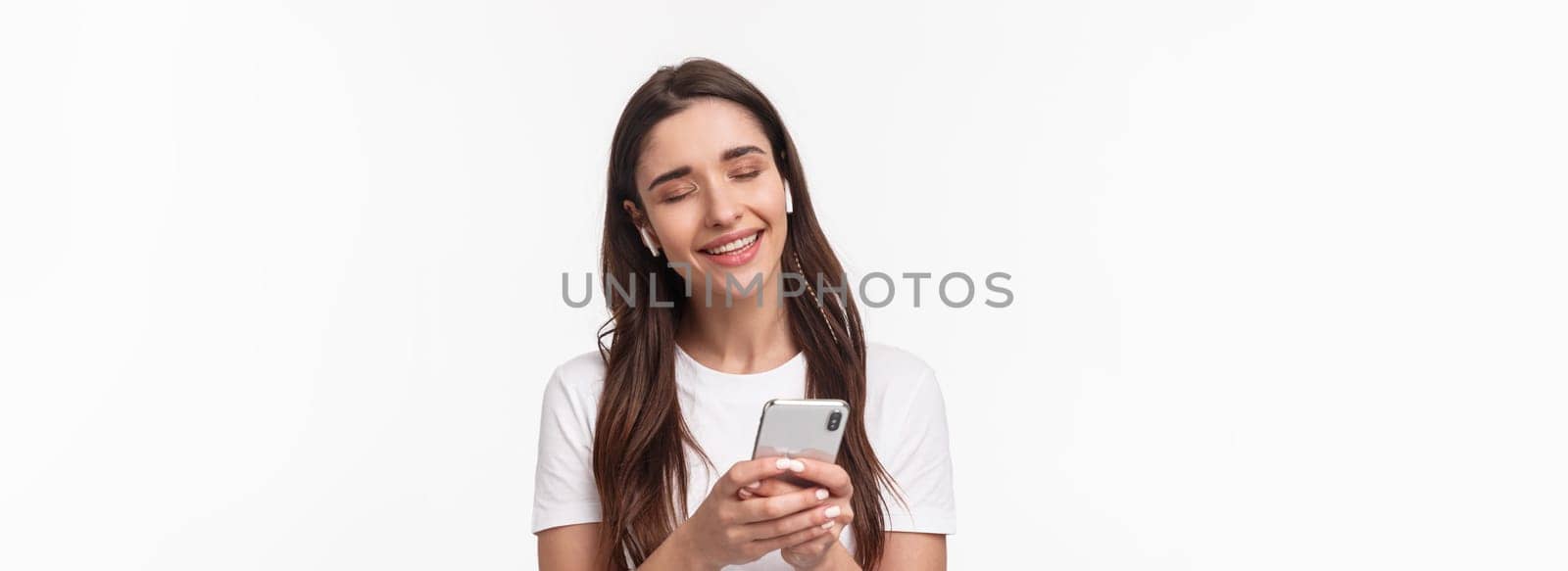 Close-up portrait of tender cute young brunette woman close eyes, shaking hand in rhythm music, smiling from delight, listening song in wireless earphones, holding smartphone.