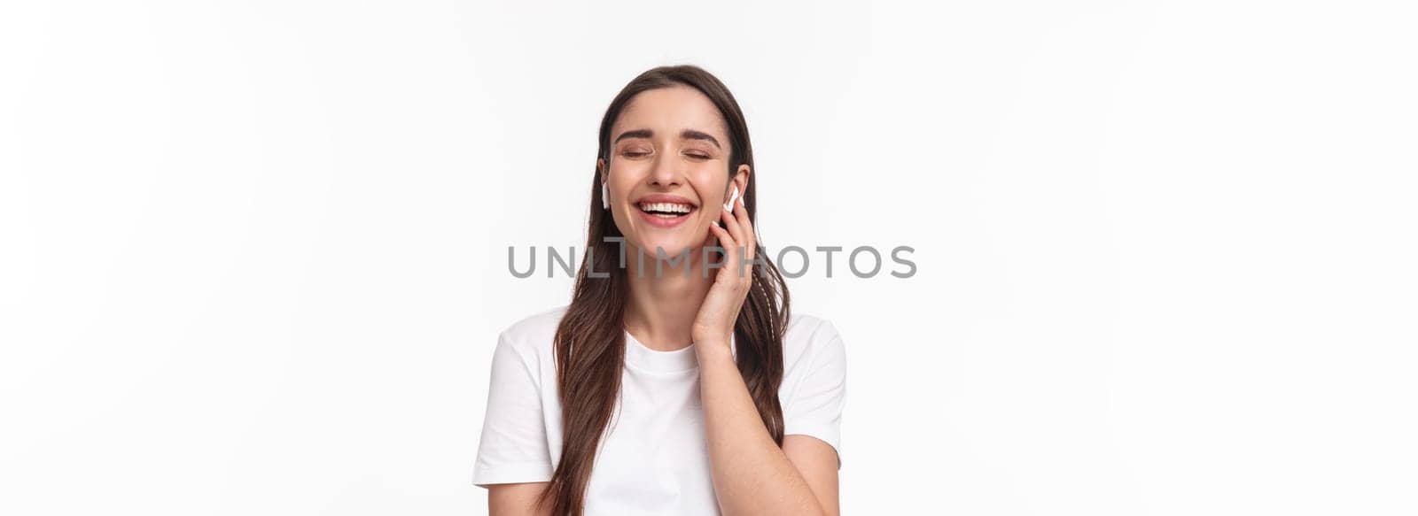 Close-up portrait of adorable caucasian woman, smiling and laughing with closed eyes, touching wireless earphones, having phone conversation with headphones, white background by Benzoix