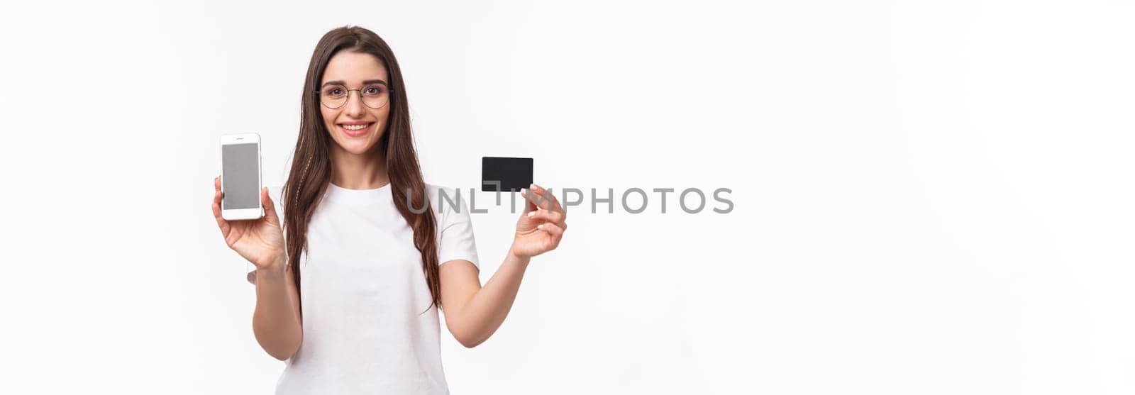 Portrait of happy friendly-looking young woman in t-shirt and glasses, showing credit card and mobile phone screen as advertise online application, internet purchase, non-cash payment.