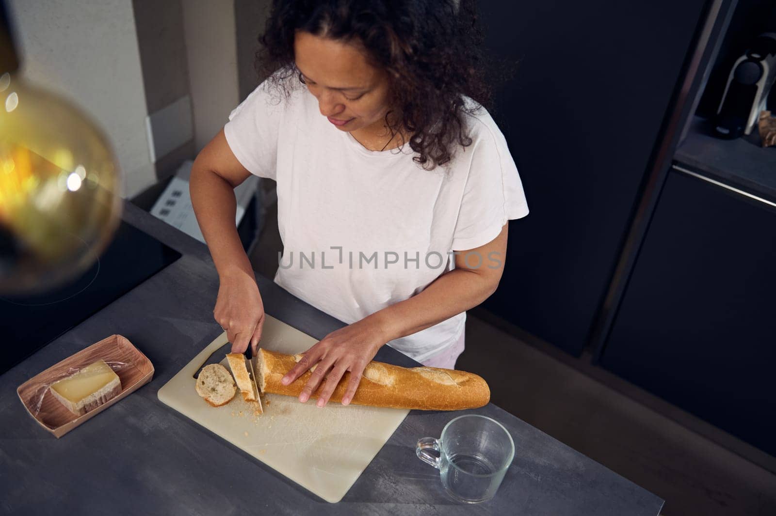 View from above of a woman using kitchen knife, cutting a loaf of whole grain French baguette bread on the cutting board, preparing delicious sandwiches with cheese for breakfast in the home kitchen.