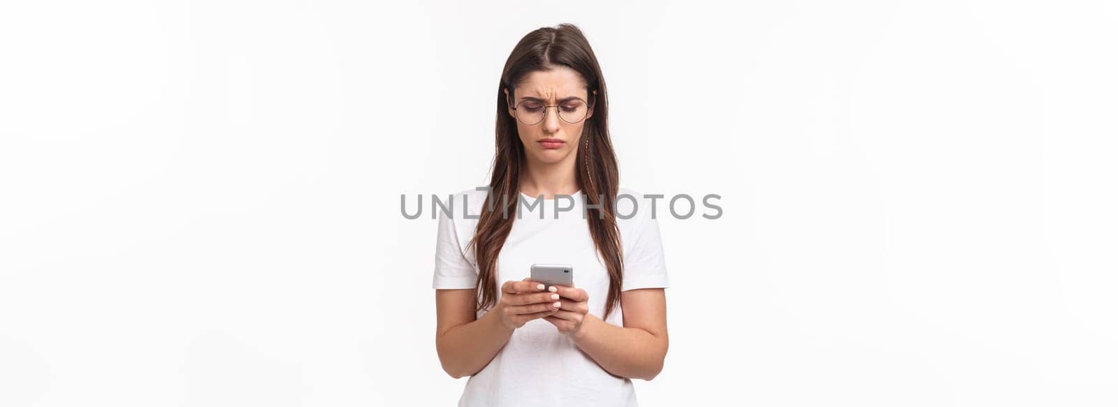 Communication, technology and lifestyle concept. Portrait of troubled and confused young woman reading strange message, frowning as looking mobile phone display, stand white background.