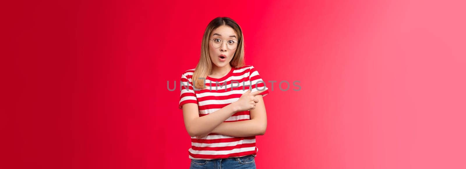 Fascinated surprised cute asian girl in glasses talking about incredible offer folding lips gasping, look wondered camera raise eyebrows astonished pointing left questioned intrigued, red background.