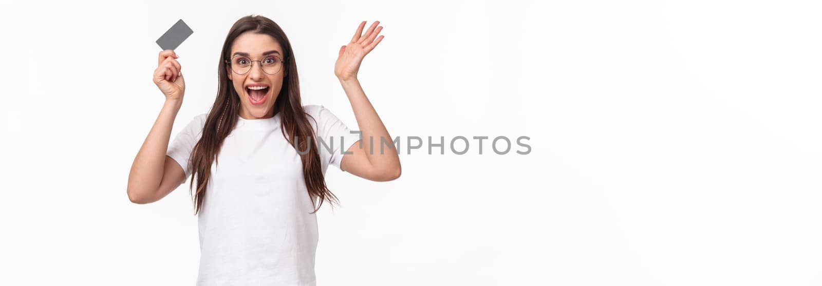 Waist-up of enthusiastic, happy surprised young woman reacting to wonderful news, raising hands up in amazement, holding credit card, receive salary, got her first paycheck, hurry to order something.