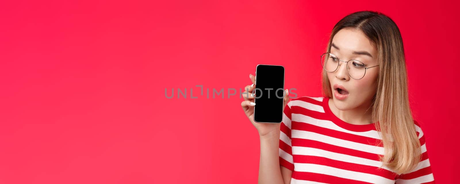 Close-up wondered surprised attractive asian blond girl wear glasses hold smartphone looking impressed amused phone screen, open mouth interesting app telephone display, stand red background.