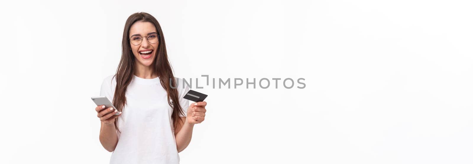 Portrait of happy smiling pretty woman in glasses holding credit card, mobile phone, laughing at camera easily paying for her purchase online store, buy things with application, white background.