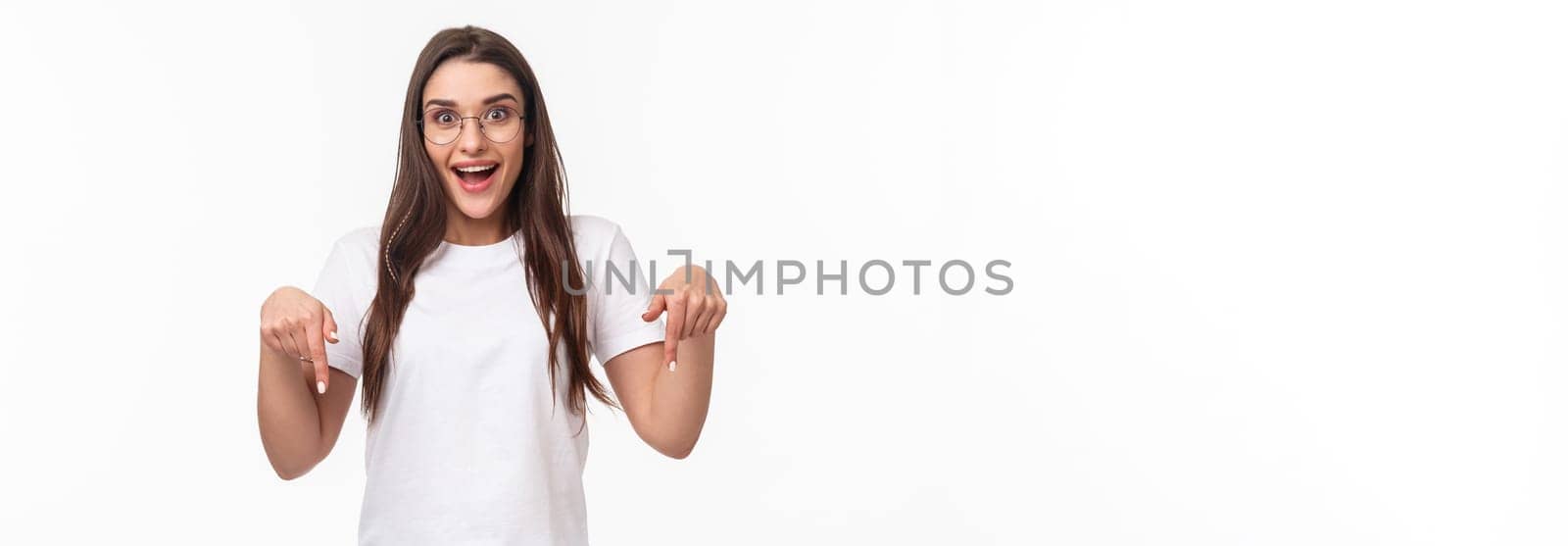 Waist-up portrait of excited young pretty woman seeing fantastic news, event promo or special spring discount, pointing fingers down and looking camera enthusiastic, stand white background.