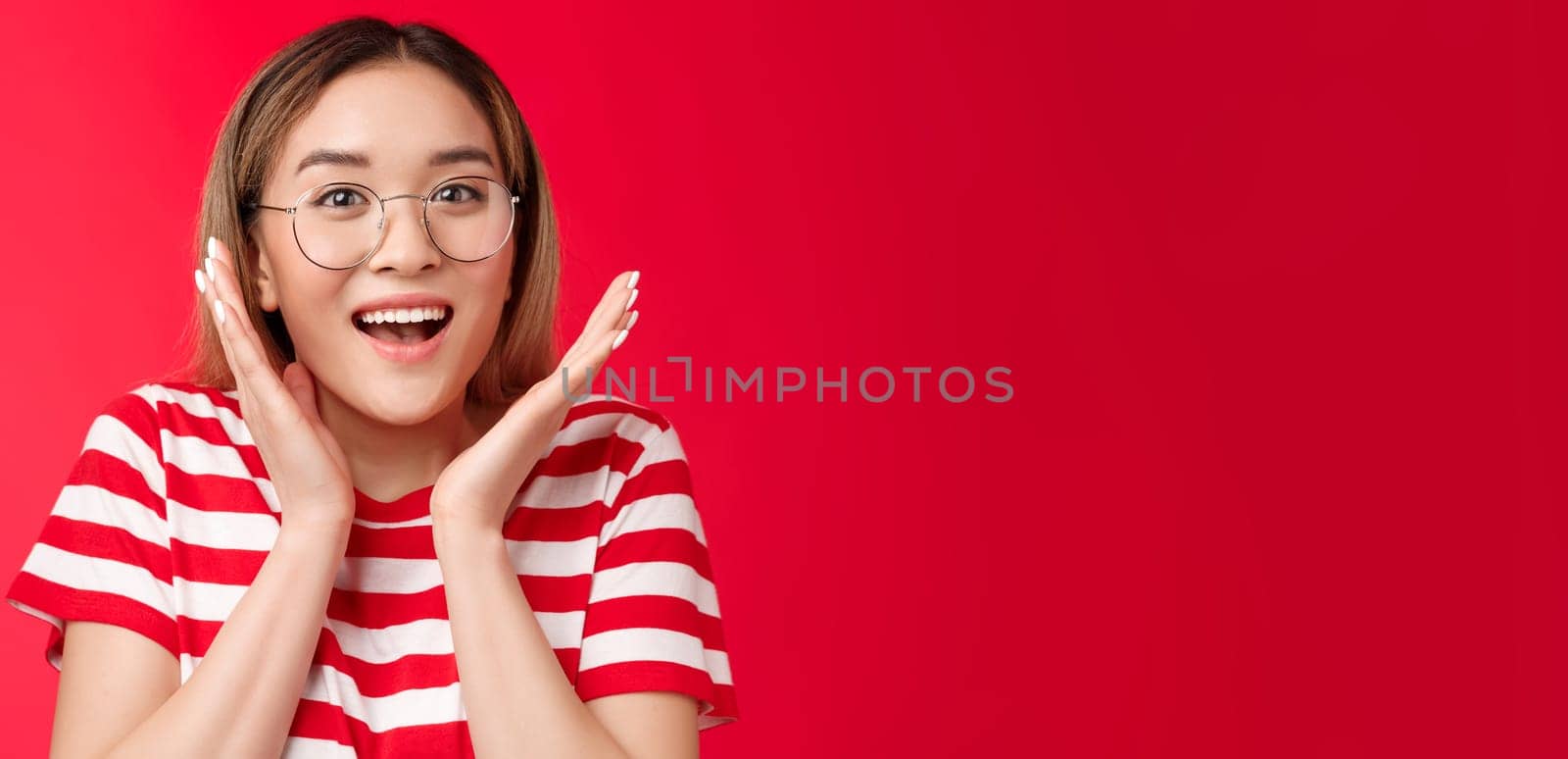 Surprised happy cheering cute asian blond girl realize got lucky smiling broadly, gesturing shaking hands excitement and joy, grinning amazed great news, stand red background celebrating win.