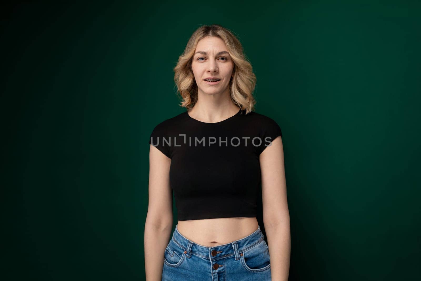 A woman is standing upright with a neutral expression on her face, positioned in front of a solid green background. She appears to be staring straight ahead with her hands resting at her sides.