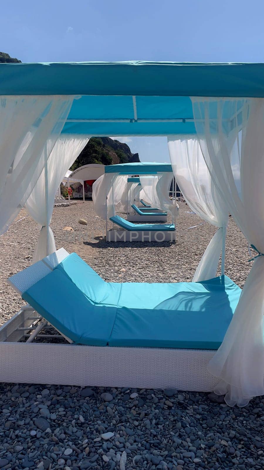 A blue and white beach umbrella with white curtains is set up on a beach. The umbrella is surrounded by several lounge chairs, creating a relaxing and inviting atmosphere