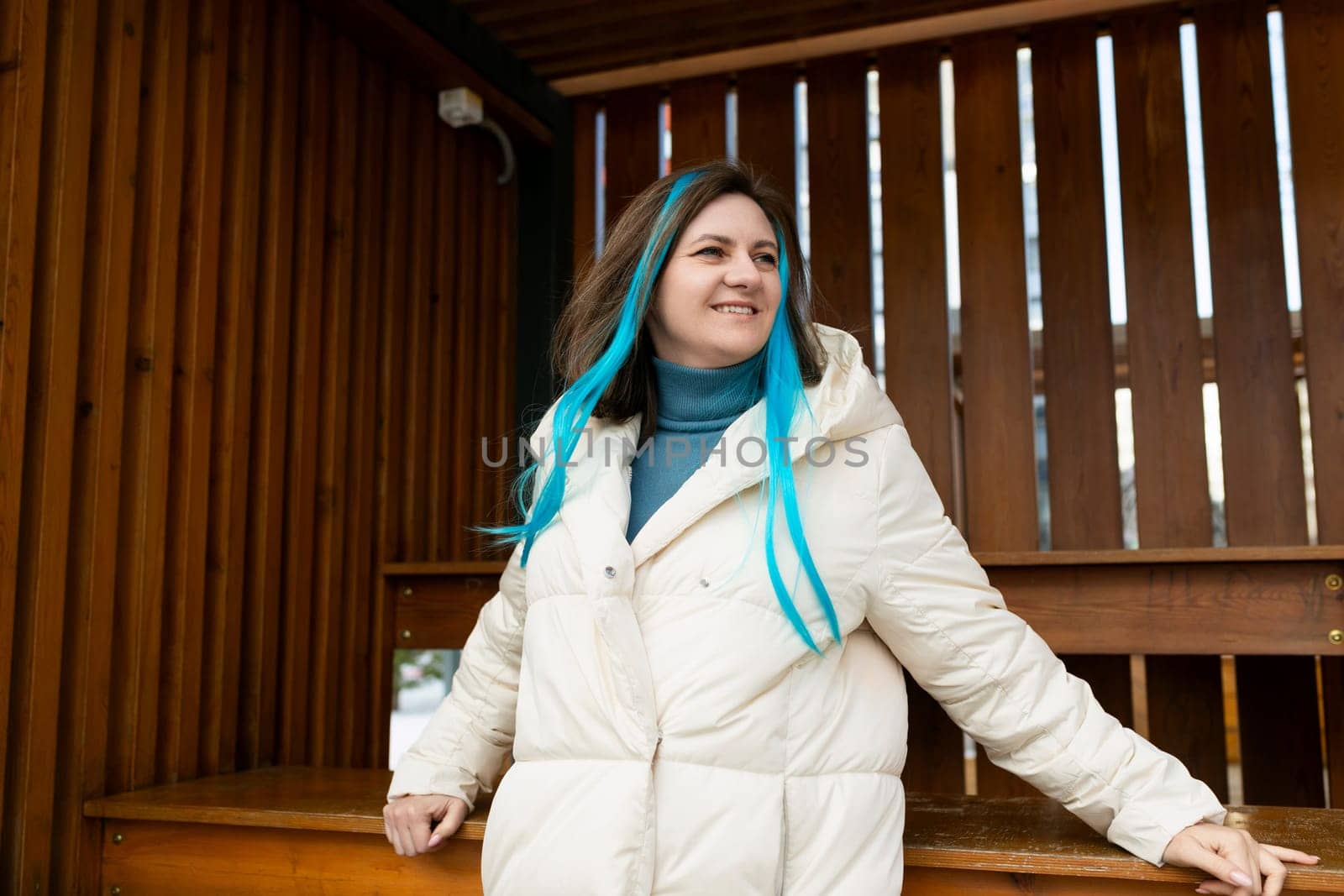 A woman wearing a white coat stands while showcasing vibrant blue hair. She exudes confidence and uniqueness with her striking appearance.