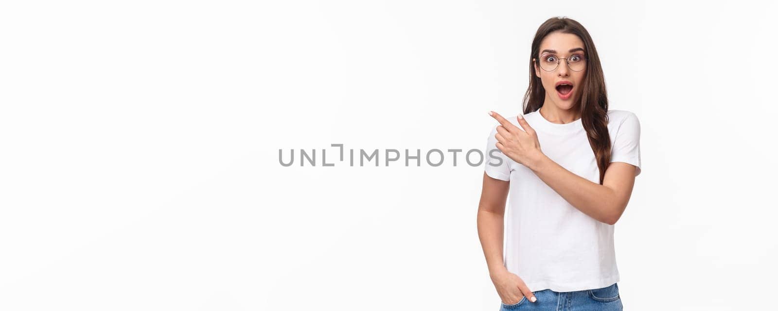 Waist-up portrait of enthusiastic, amazed and impressed young attractive girl in glasses talking about amazing new event or promo she saw online, pointing finger upper left corner, gasping astonished.