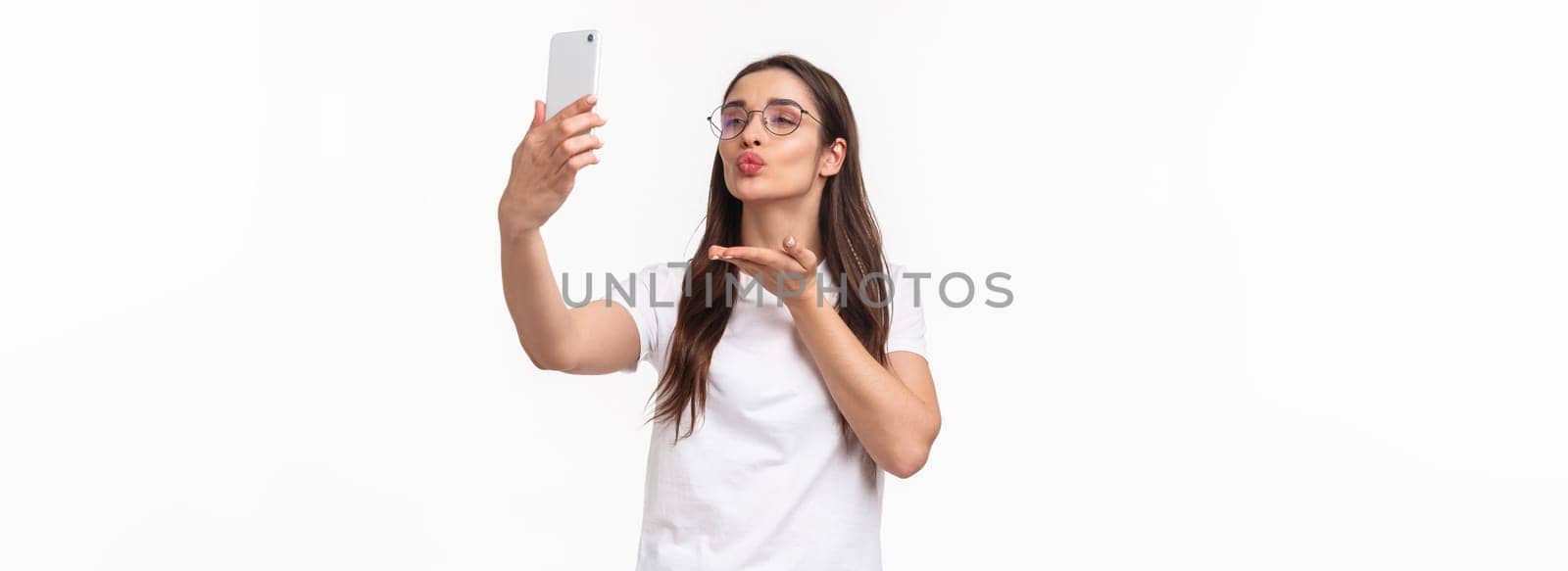 Communication, technology and lifestyle concept. Lovely attractive woman in glasses, sending air kiss to her followers online live stream with app, taking photo with filters, hold mobile phone.