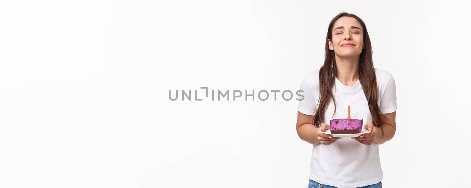 Entertainment, fun and holidays concept. Portrait of romantic beautiful young b-day girl celebrating throwing party, holding piece of birthday cake with candle, making wish with smile and closed eyes.