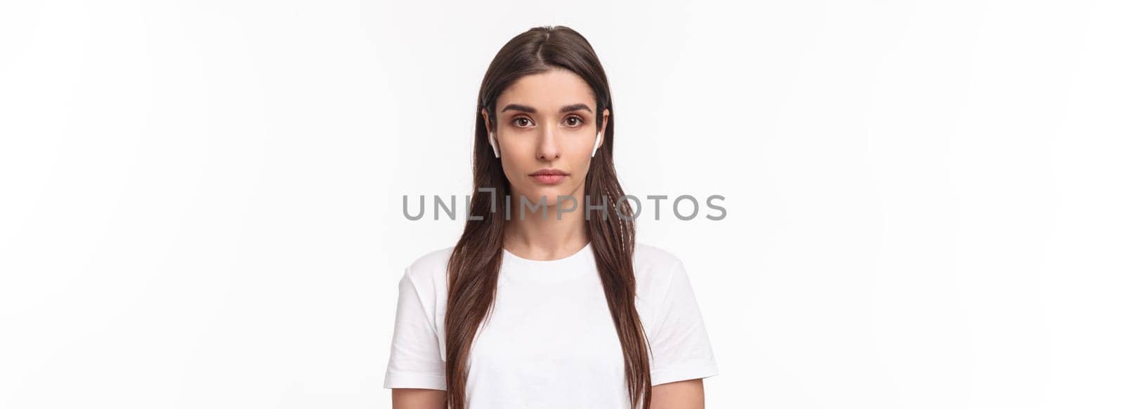 Close-up portrait of serious-looking attractive caucasian woman in wireless earphones, looking at camera with relaxed no emotions face, standing white background. Copy space