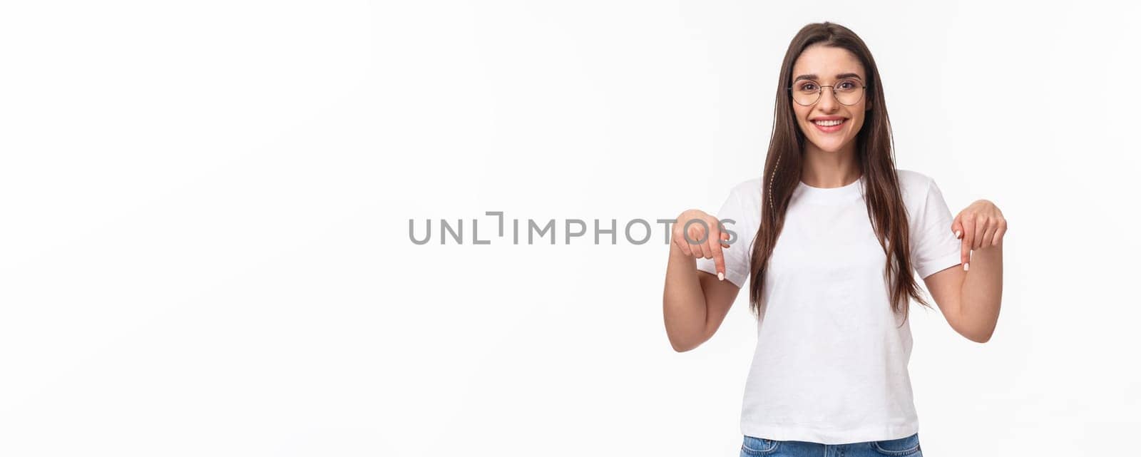 Click here. Portrait of brunette gorgeous young woman in glasses, pointing fingers down to show advertisement, smiling pleased, suggest visit link, recommend download or subscribe, white background.