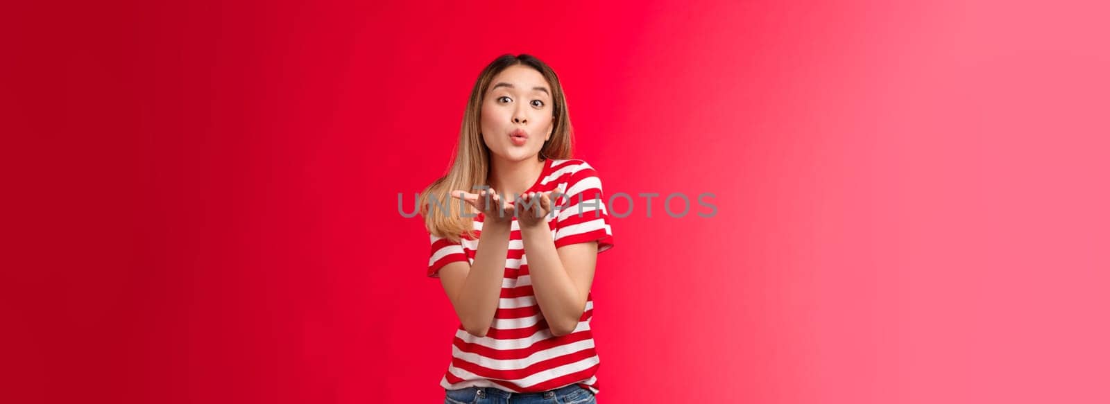 Silly glamour tender asian blond modern girl send you sweet kisses. Joyful urban woman wear striped t-shirt hold palms near fold lips blow kiss air mwah, stand red background glamour.