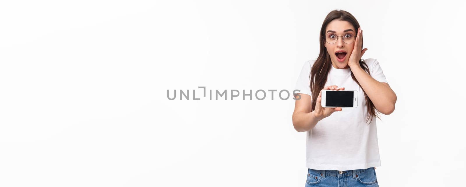 Communication, technology and lifestyle concept. Portrait of surpirsed and impressed beautiful young girl in glasses, gasping amazed touch cheek stunned, showing cool content on mobile phone display.