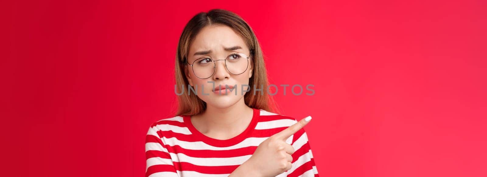 Disappointed doubtful frowning asian girl asking question reassure, looking pointing left displeased, unimpressed with strange place, dislike interiour sulking distressed, stand red background.