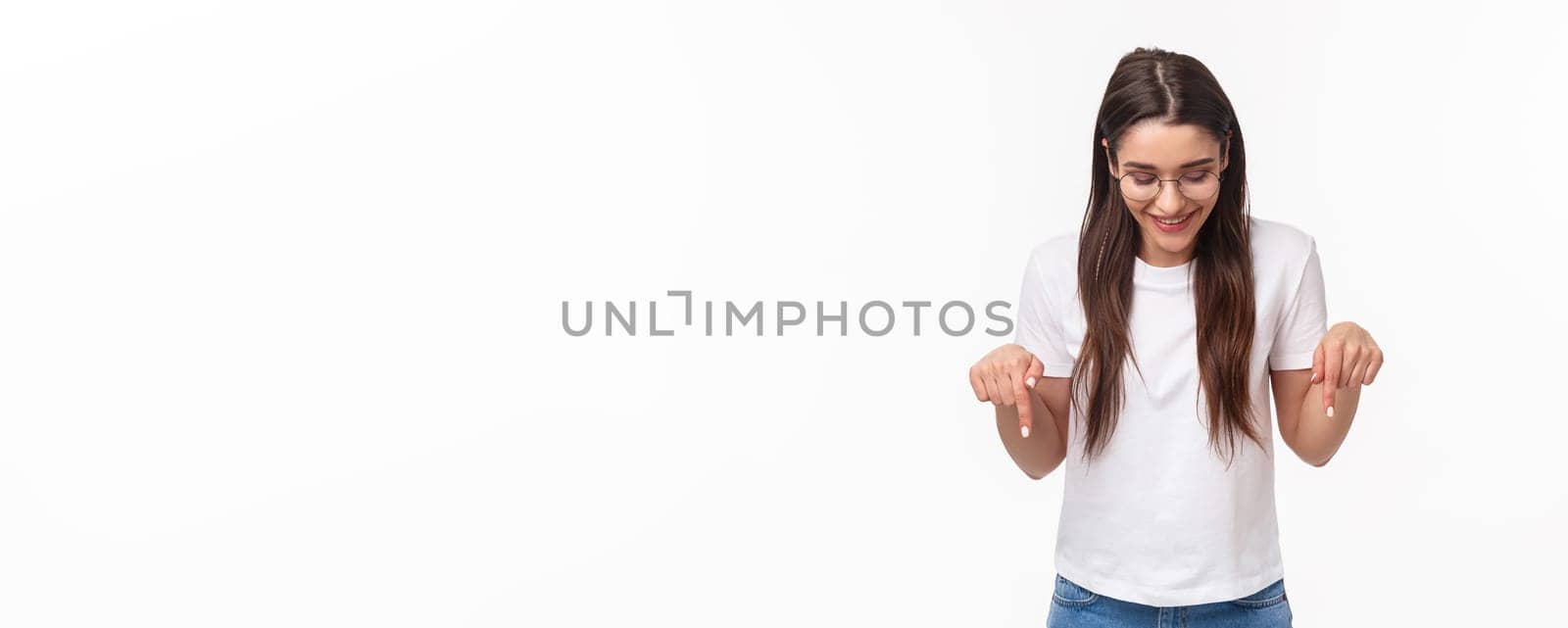 Waist-up portrait of enthusiastic, smiling happy brunette female student in glasses and t-shirt, looking pointing down, bottom advertisement, glancing pleased, standing white background.