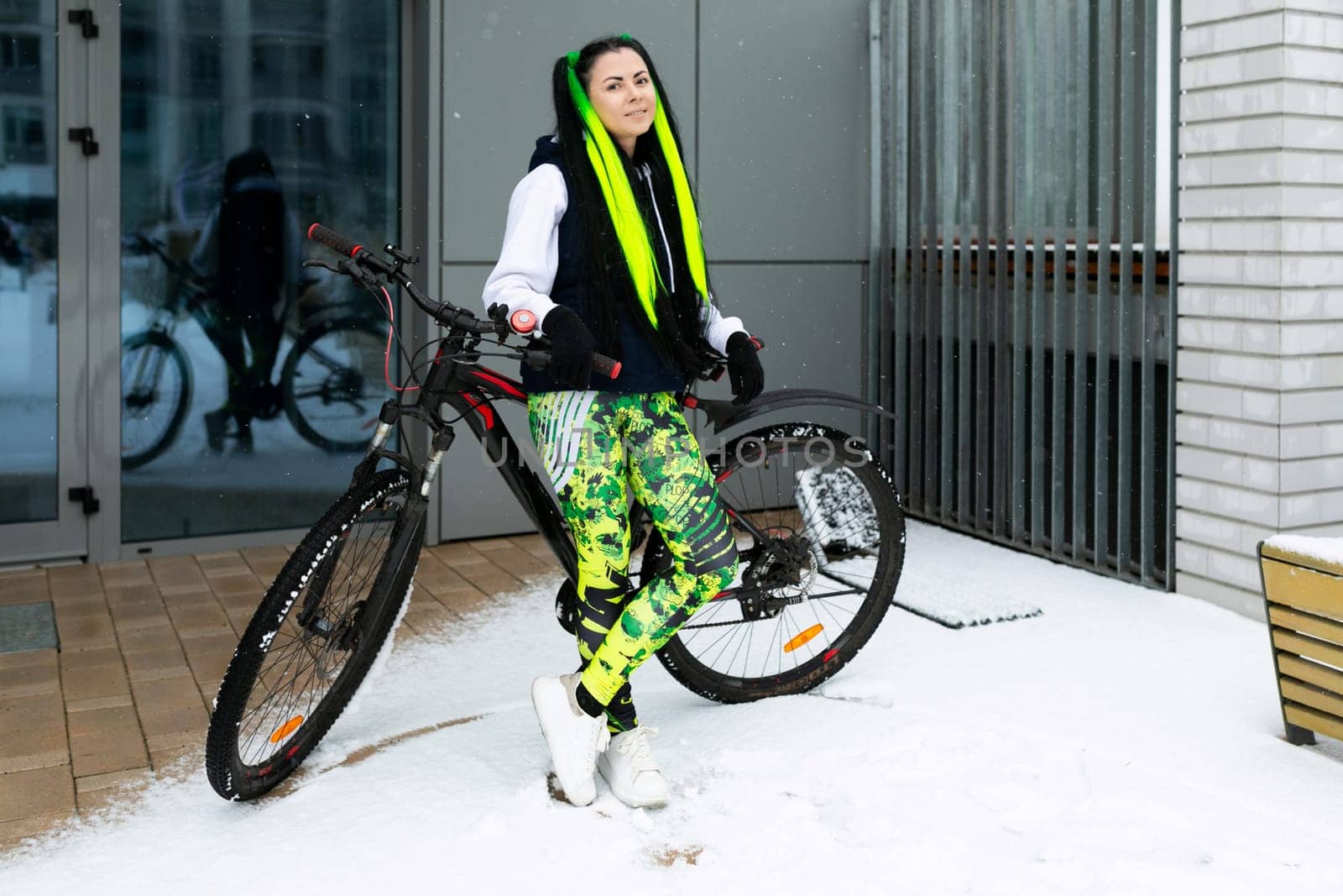 A woman standing next to a bike in a snowy landscape. She is wearing warm clothing, and there are footprints in the snow. The bike is partially covered in snow.