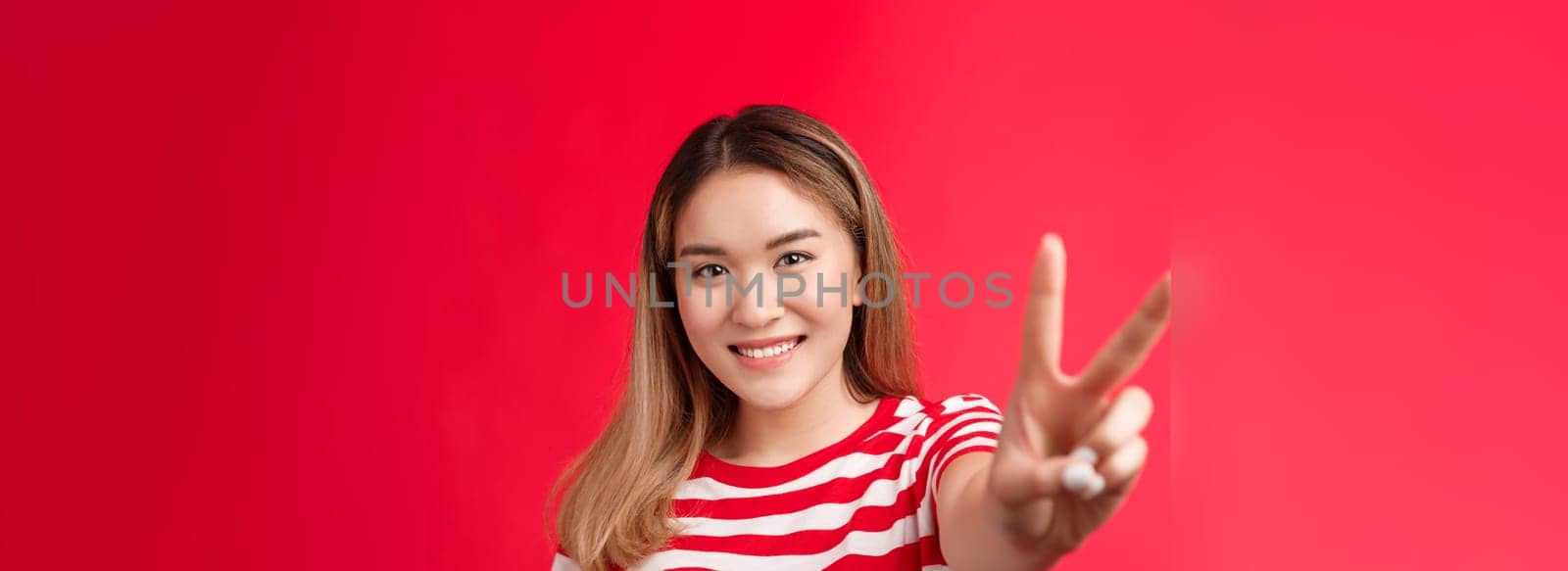 Optimsitic positive young asian female, support lgbt community, pull hand forward show peace victory sign, smiling happily feel lucky, stand red background joyful, enjoy summer vacation.