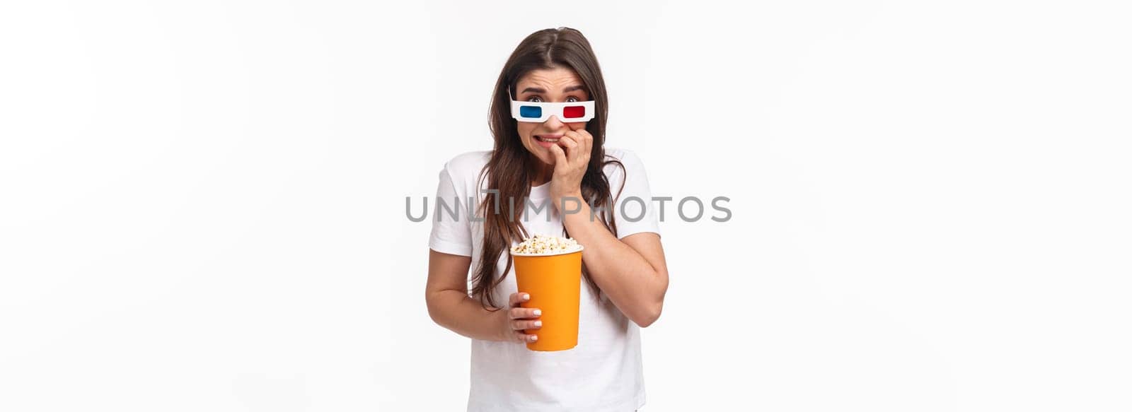 Entertainment, fun and holidays concept. Portrait of scared young cute girl in 3d glasses, eating popcorn, stooping and biting fingers as being scared of horror movie watching in cinema by Benzoix