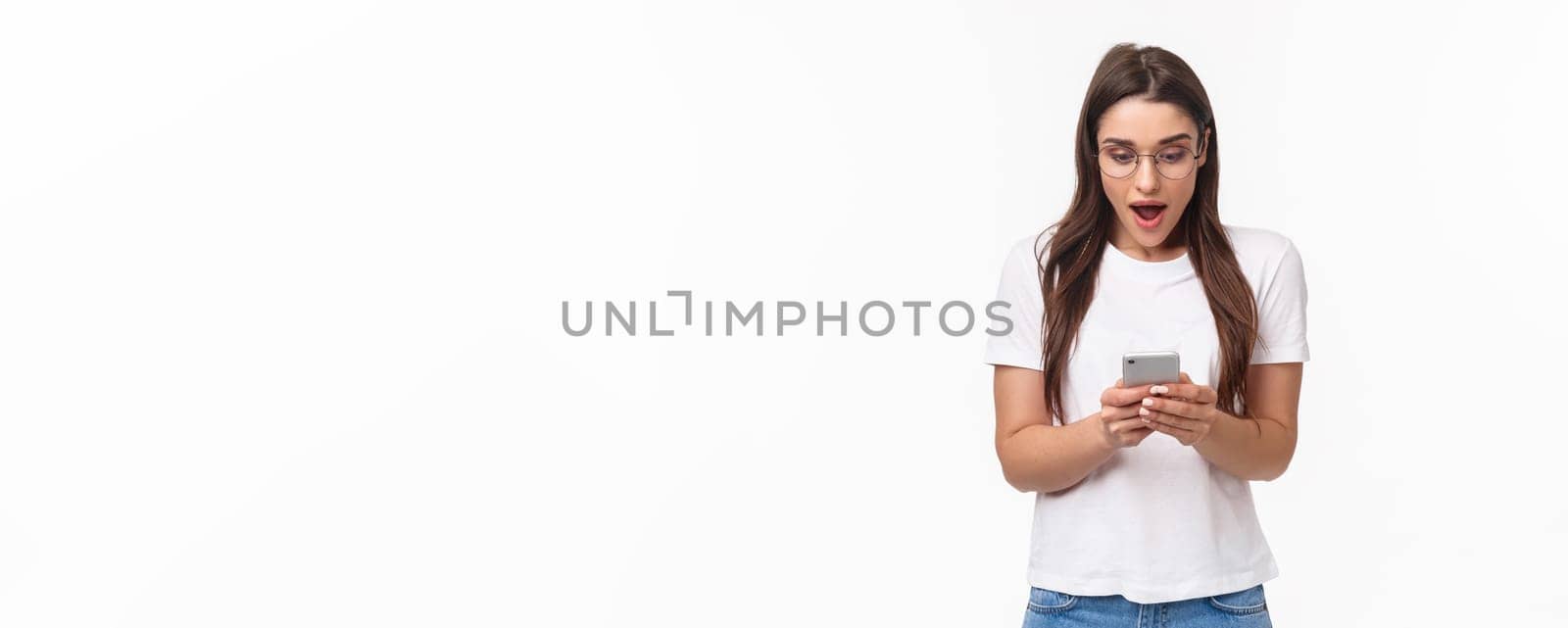 Communication, technology and lifestyle concept. Portrait of surprised and amazed, excited young caucasian woman in glasses, looking at mobile phone, receive awesome new via text message by Benzoix