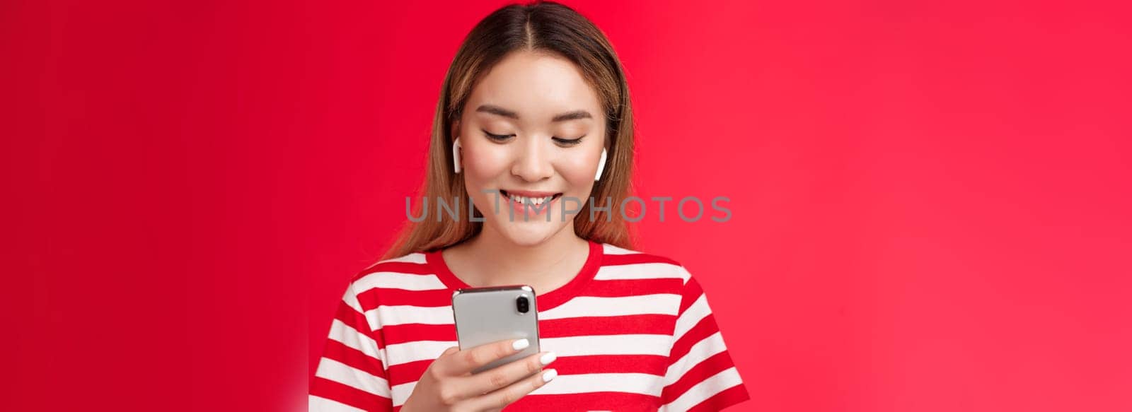 Carefree modern asian blond girl picking song wear wireless earphones, listen music using online song platform app, hold smartphone look pleased smiling phone screen, stand red background.