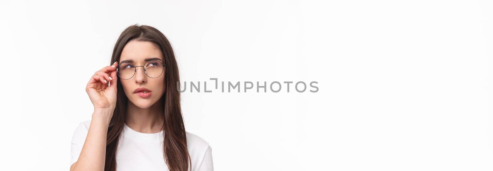 Close-up portrait of smart and thoughtful beautiful young girl in glasses, thinking, squinting suspicious and biting lip look up while pondering something, have interesting idea, white background.