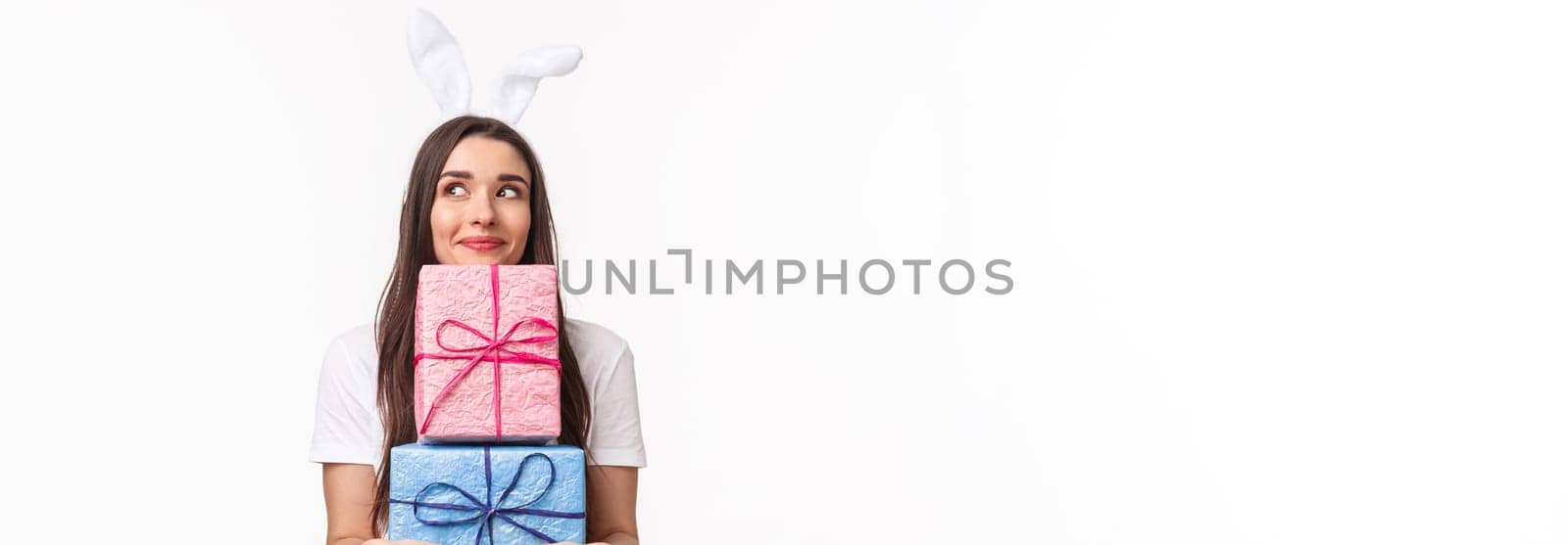 Celebration, holidays and presents concept. Portrait of silly and cute, lovely young girl in rabbit ears, holding presents, daydreaming, smiling and looking away, giving out gift, white background by Benzoix