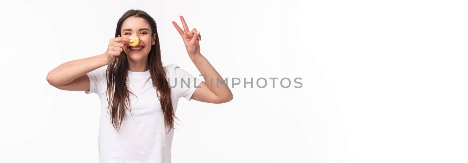Portrait of positive, cheerful young girl eating sweets, holding macaron over nose, make peace sign, look kawaii and silly, laughing joyful, visit favorite dessert cafe, white background.