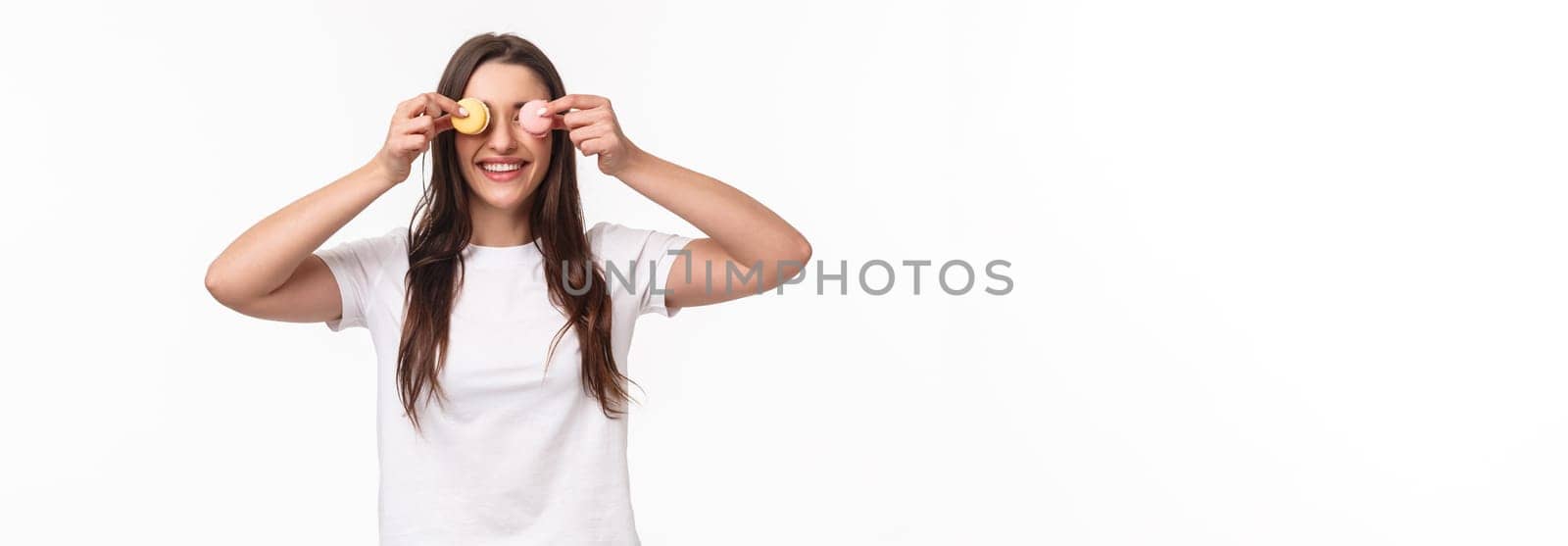 Waist-up portrait of funny and cute, adorable young woman holding two desserts macarons over eyes and smiling, playing with food, fool around, standing upbeat over white background.