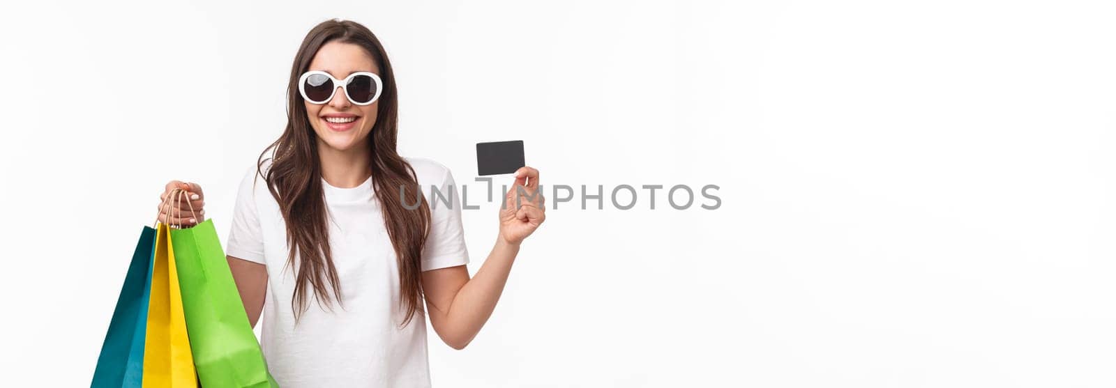 Shopping, leisure and lifestyle concept. Portrait of carefree, relaxed young woman spending her money on purchasing new clothes, products, holding shop bags and showing credit card, smiling pleased.