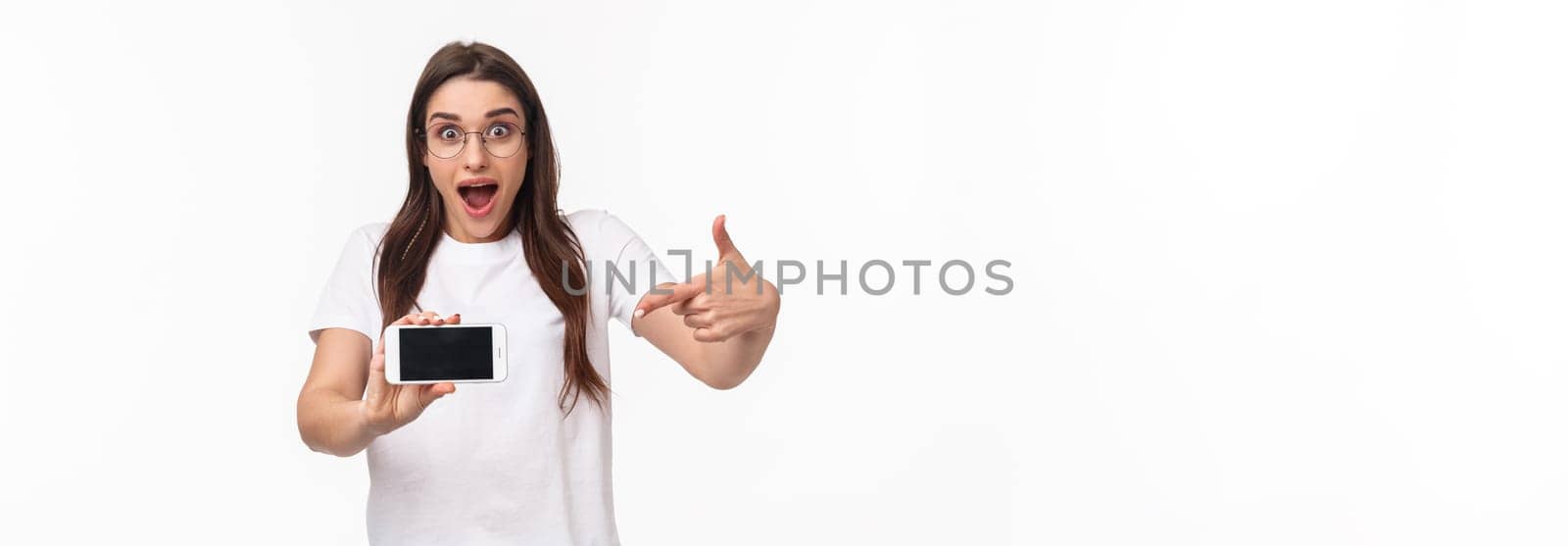 Communication, technology and lifestyle concept. Portrait of surprised and excited young woman in glasses hurry up with announcement of awesome new application, pointing at mobile phone.