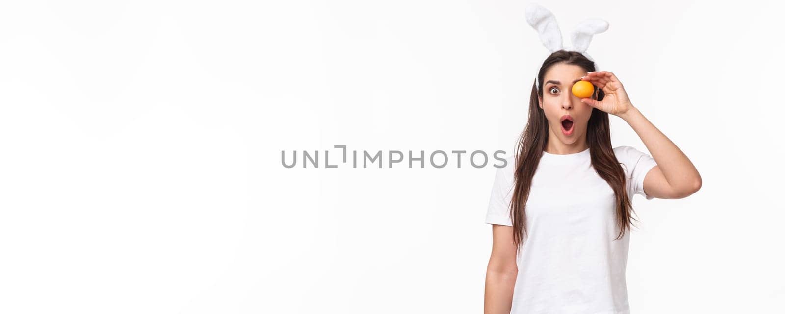 Waist-up portrait of amazed and surprised young woman seeing something awesome, hold colored egg over eye, staring shocked camera, gasping impressed, wear rabbit ears, celebrate Easter day.