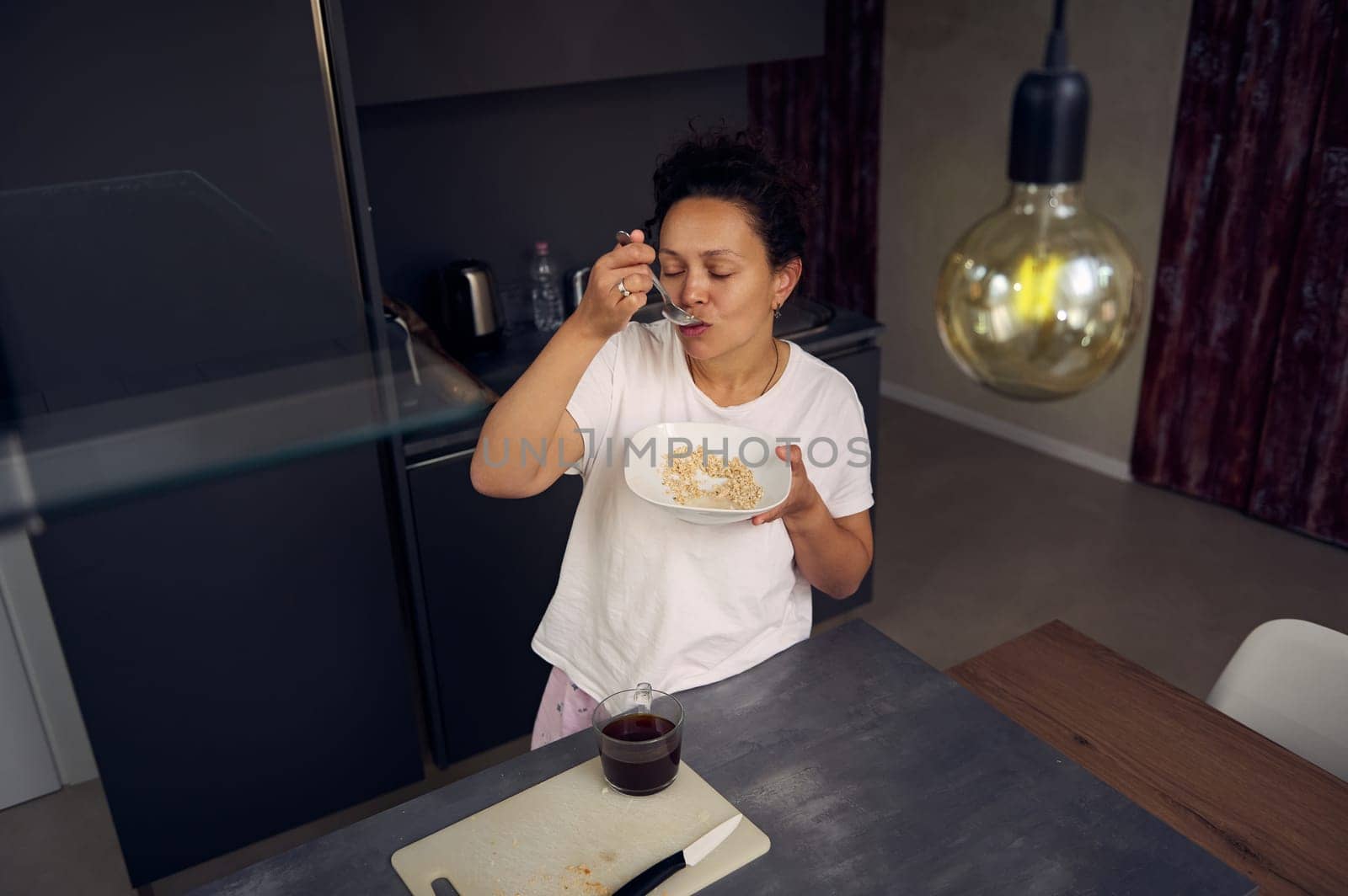 Latin American young woman 40s, smiling, drinking coffee and eating healthy muesli for breakfast in the morning at home kitchen, dressed in white pajamas. Start your day with a cup of caffeine drink