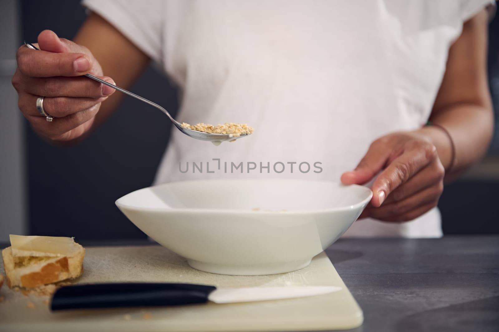 Details on the hand of woman in white t-shirt, holding a spoon with wholegrain muesli granola above a white bowl, enjoying a healthy breakfast in the morning. Healthy eating, slimming and diet concept