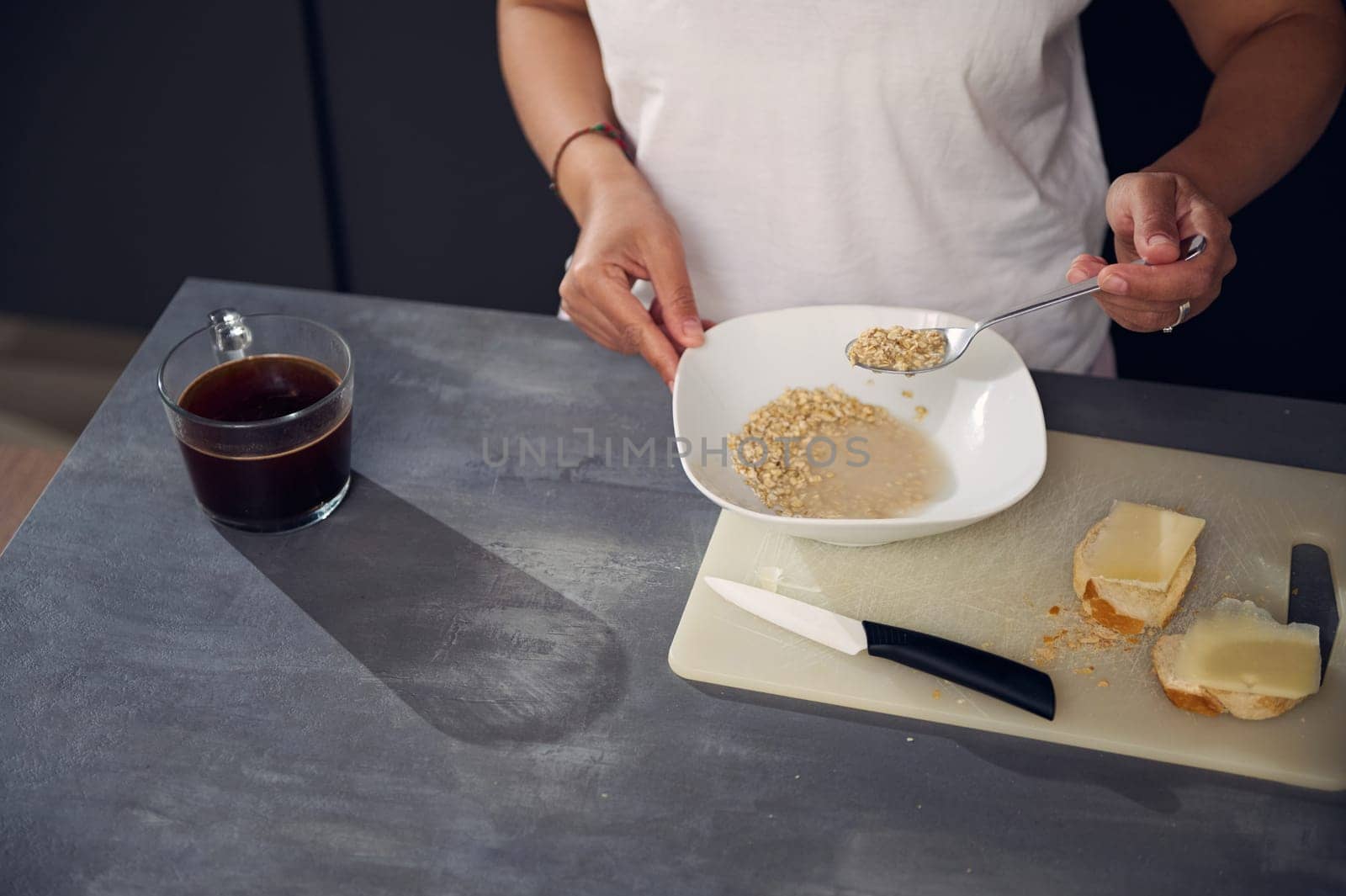 View from above of female in white t-shirt, holding a spoon with wholegrain muesli granola above a white bowl, enjoying a healthy breakfast in the morning. Healthy eating, slimming and diet concept