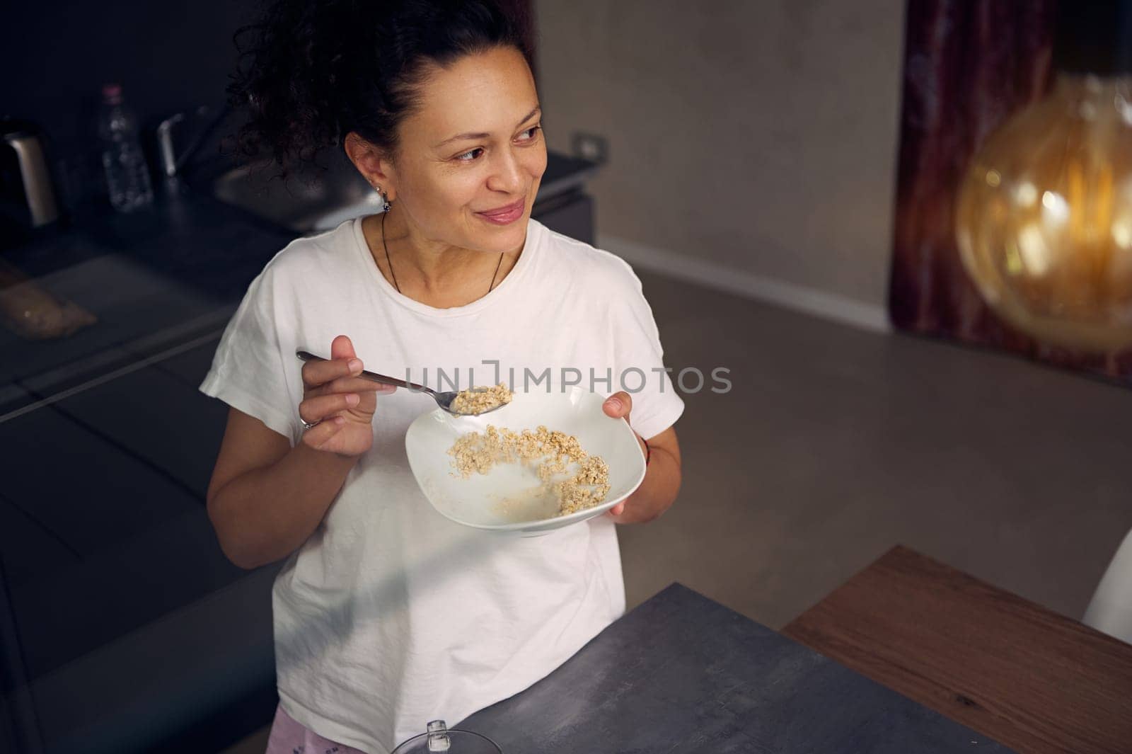 Young beautiful multi ethnic woman in pajamas, smiling looking aside, standing at kitchen table and having a healthy breakfast before work. People. Healthy eating concept. Food and drink consumerism