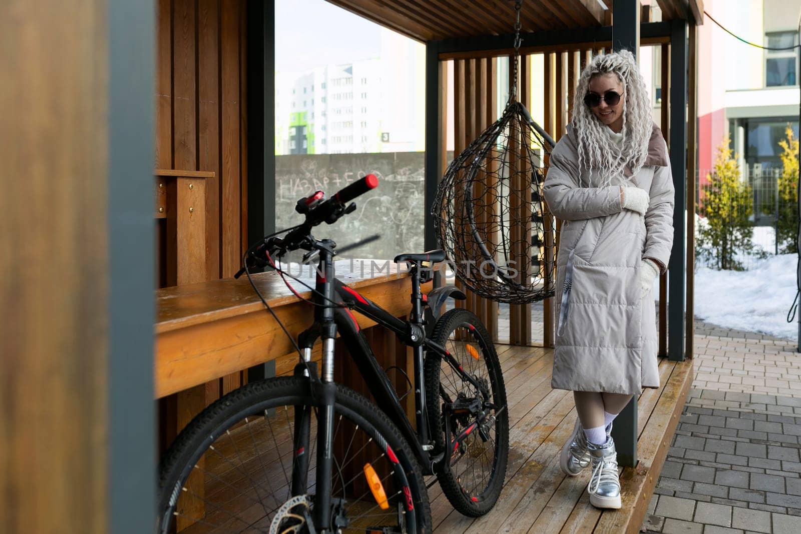 A woman wearing casual clothing is standing next to a bicycle on a wooden platform. The bike has a basket, and the woman appears to be adjusting something on it. The background shows trees and a clear sky.