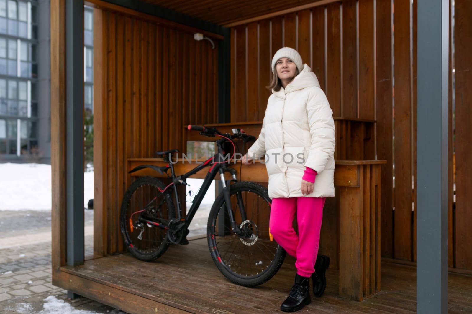 A woman wearing a white jacket is standing next to a bike, looking towards the camera. The bike is parked on a sidewalk, with buildings in the background.